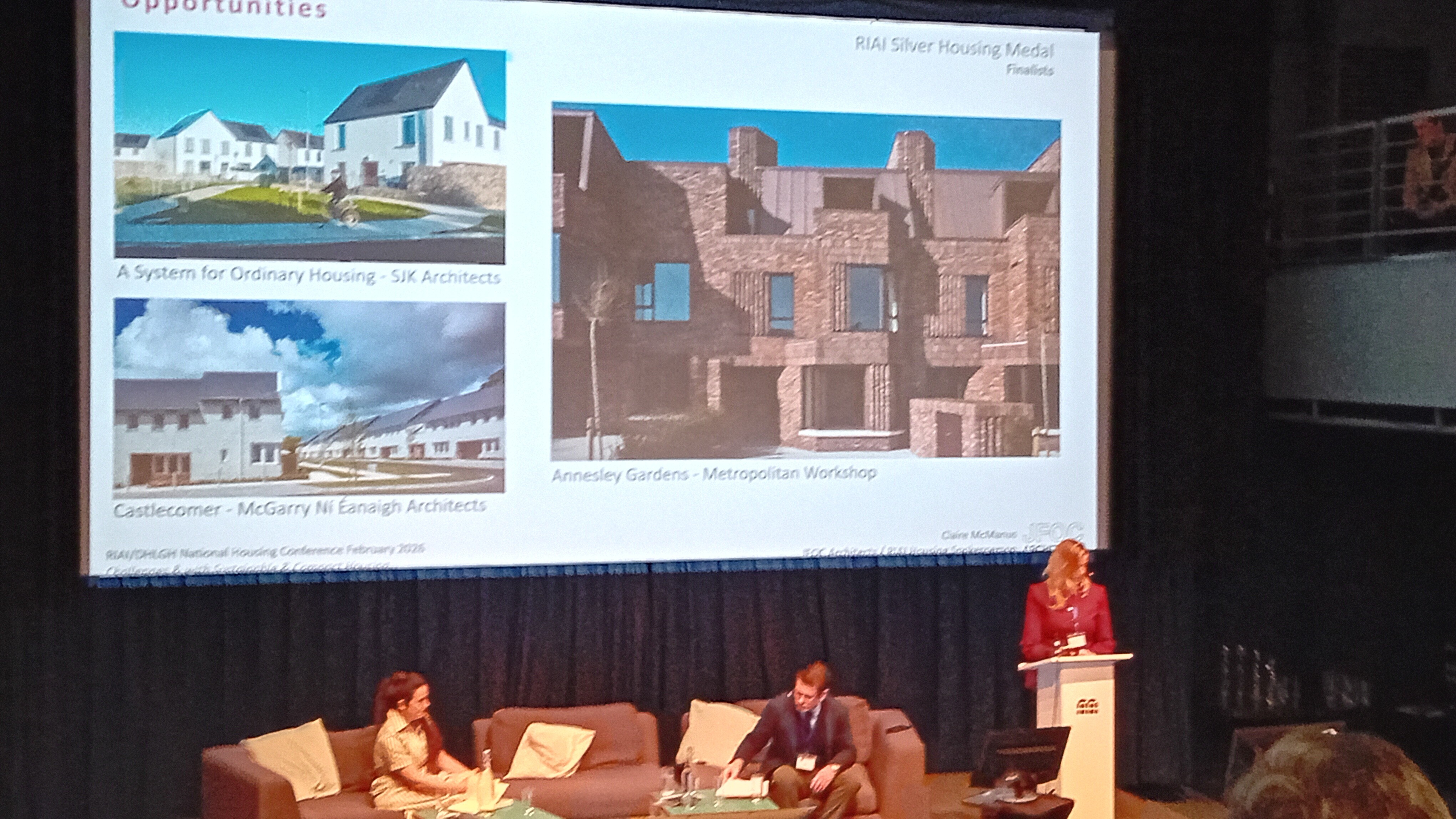 Fíonnadh McGonigle and colleagues sitting on the stage at the Housing Conference, there is a woman speaking at the podium and a presentation on a large screen