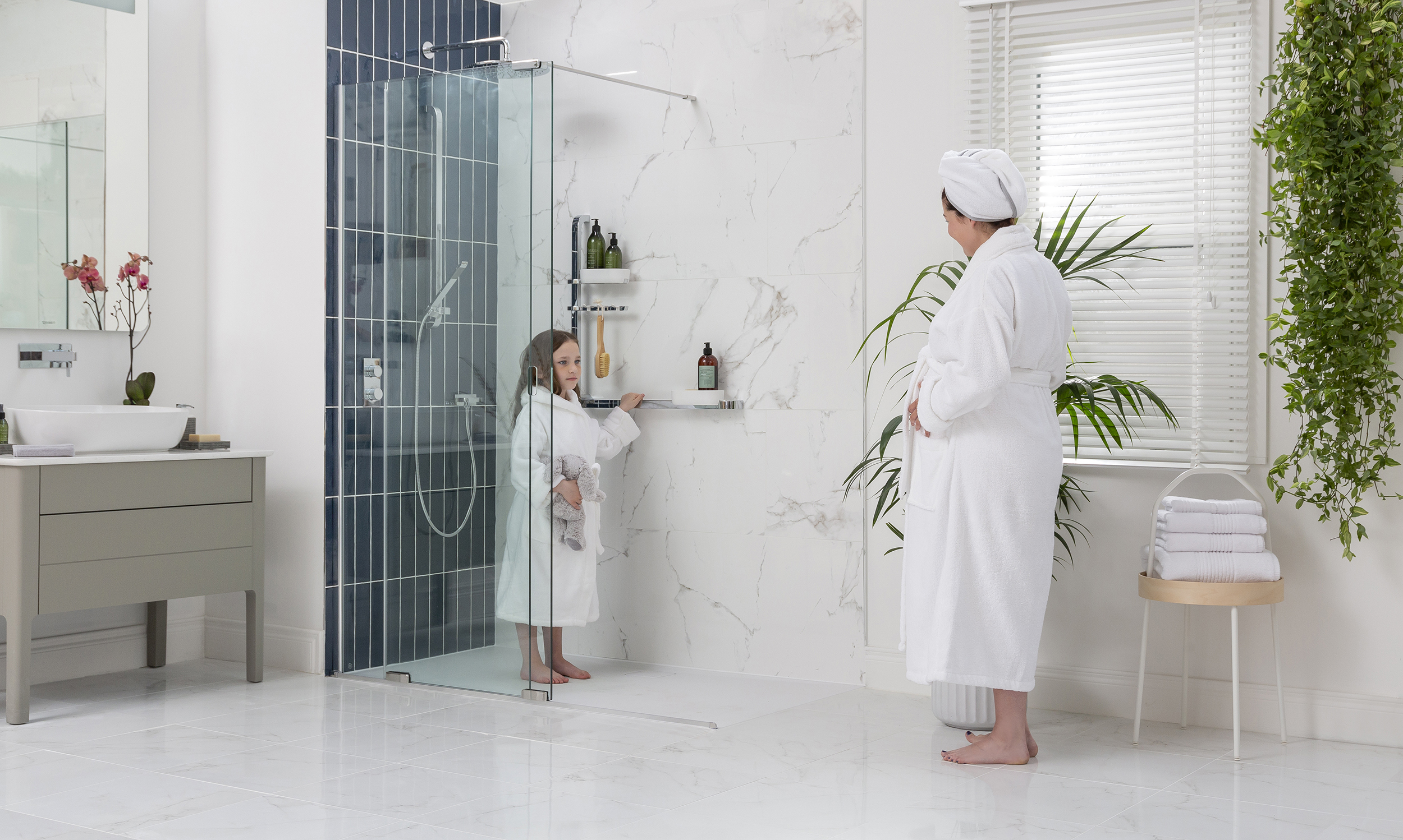 Photograph of a bathroom with a women and young child standing in their bathrobes. The child is standing in the walk in shower and beside her on the wall is a Merlyn revo shower rail