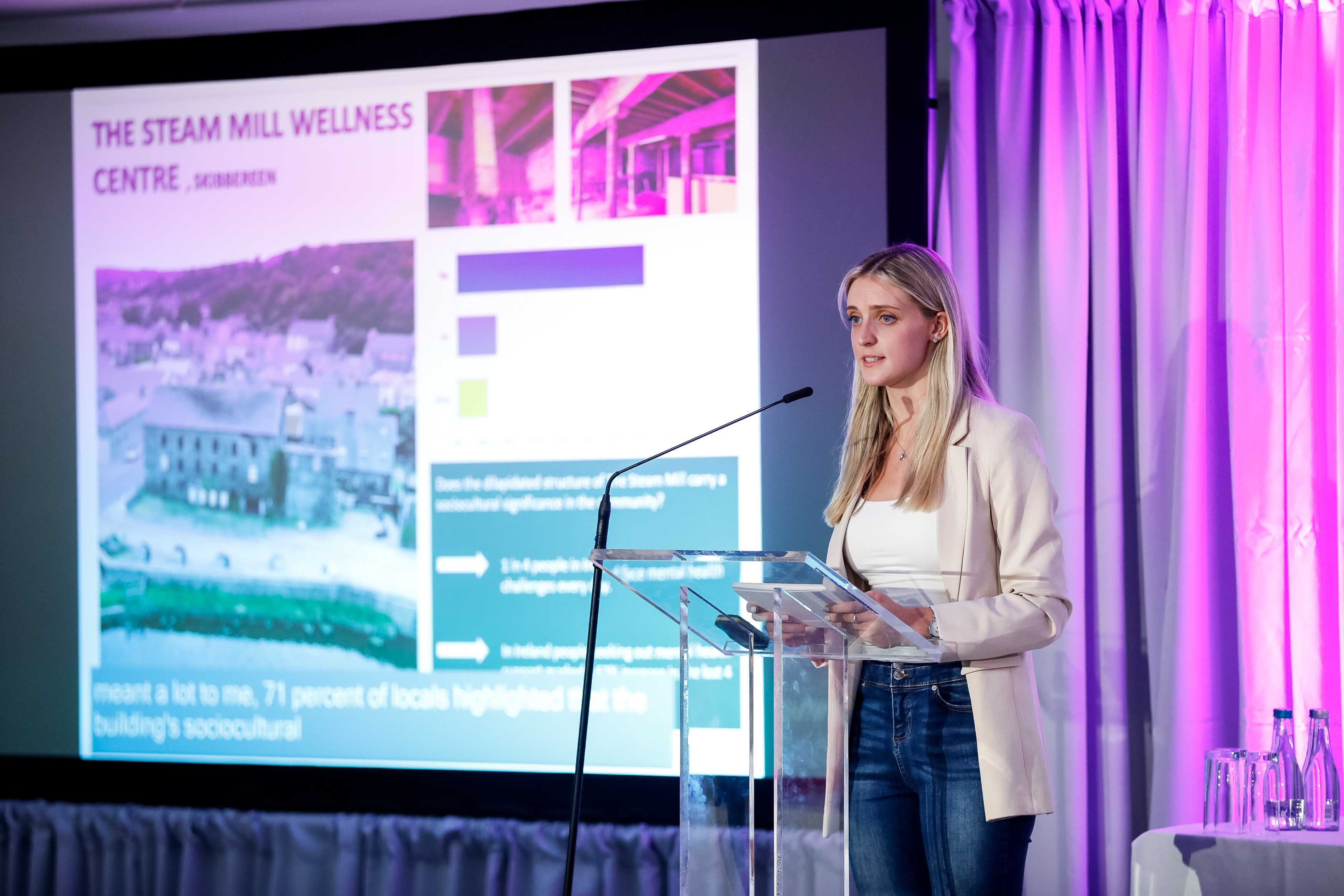Photograph of young woman speaking at a podium with cue cards in hand and a slideshow running in the background